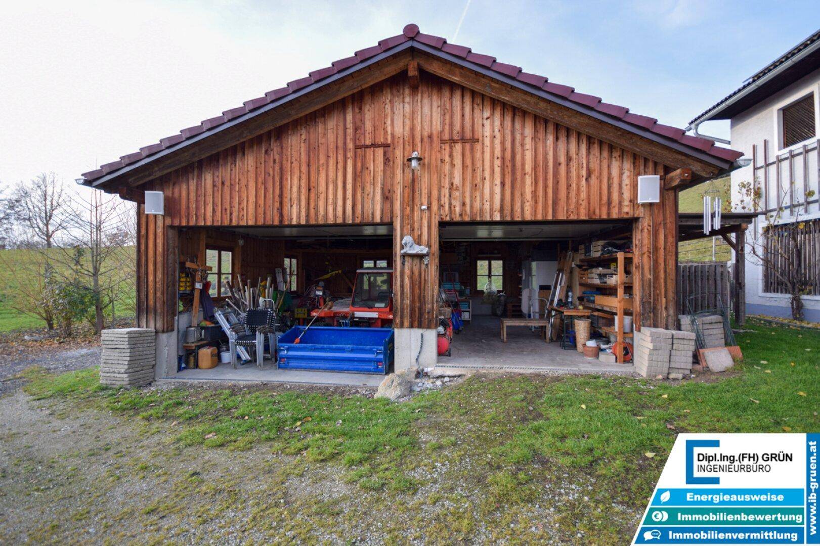 Offene Doppelgarage aus Holz mit Werkzeugen und Geräten, umgeben von ländlicher Landschaft.