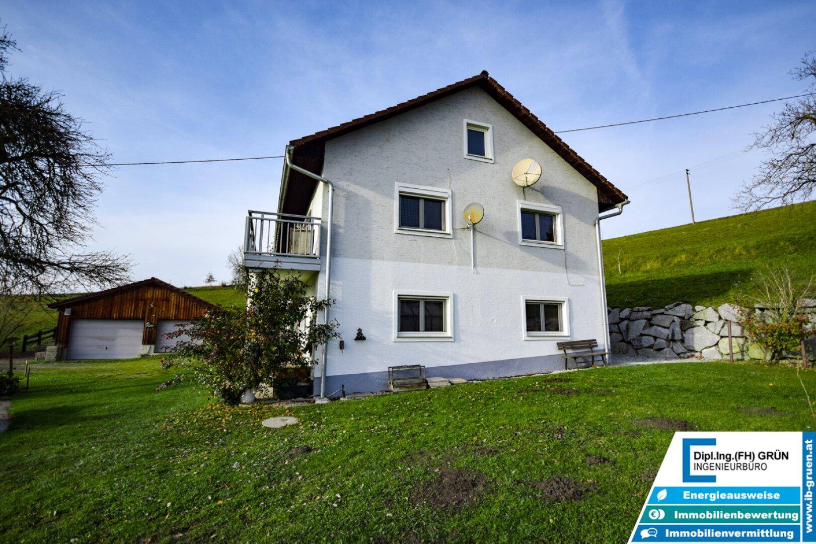 Ländliches Wohnhaus mit weißer Fassade, Balkon und Satteldach, eingebettet in eine grüne Hügellandschaft.
