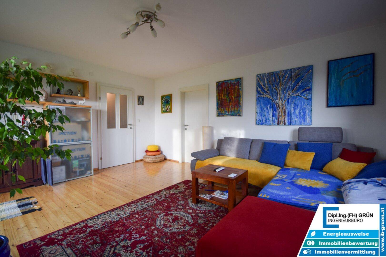 Helles Wohnzimmer mit Holzboden, großem Ecksofa und farbenfrohen Kunstwerken an den weißen Wänden.