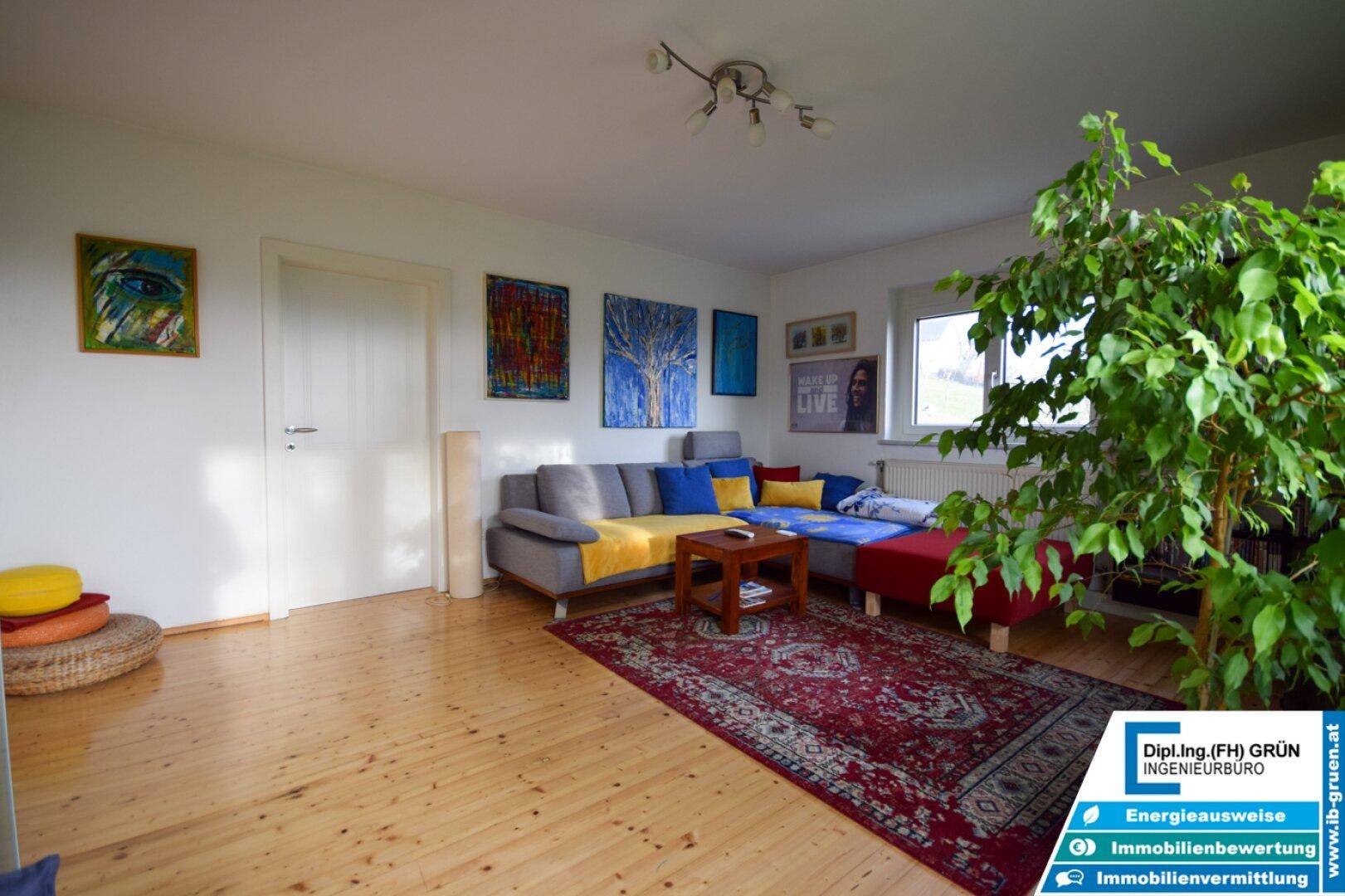 Geräumiges Wohnzimmer mit Holzboden, großem Fenster und Blick ins Grüne, dekoriert mit bunten Bildern.
