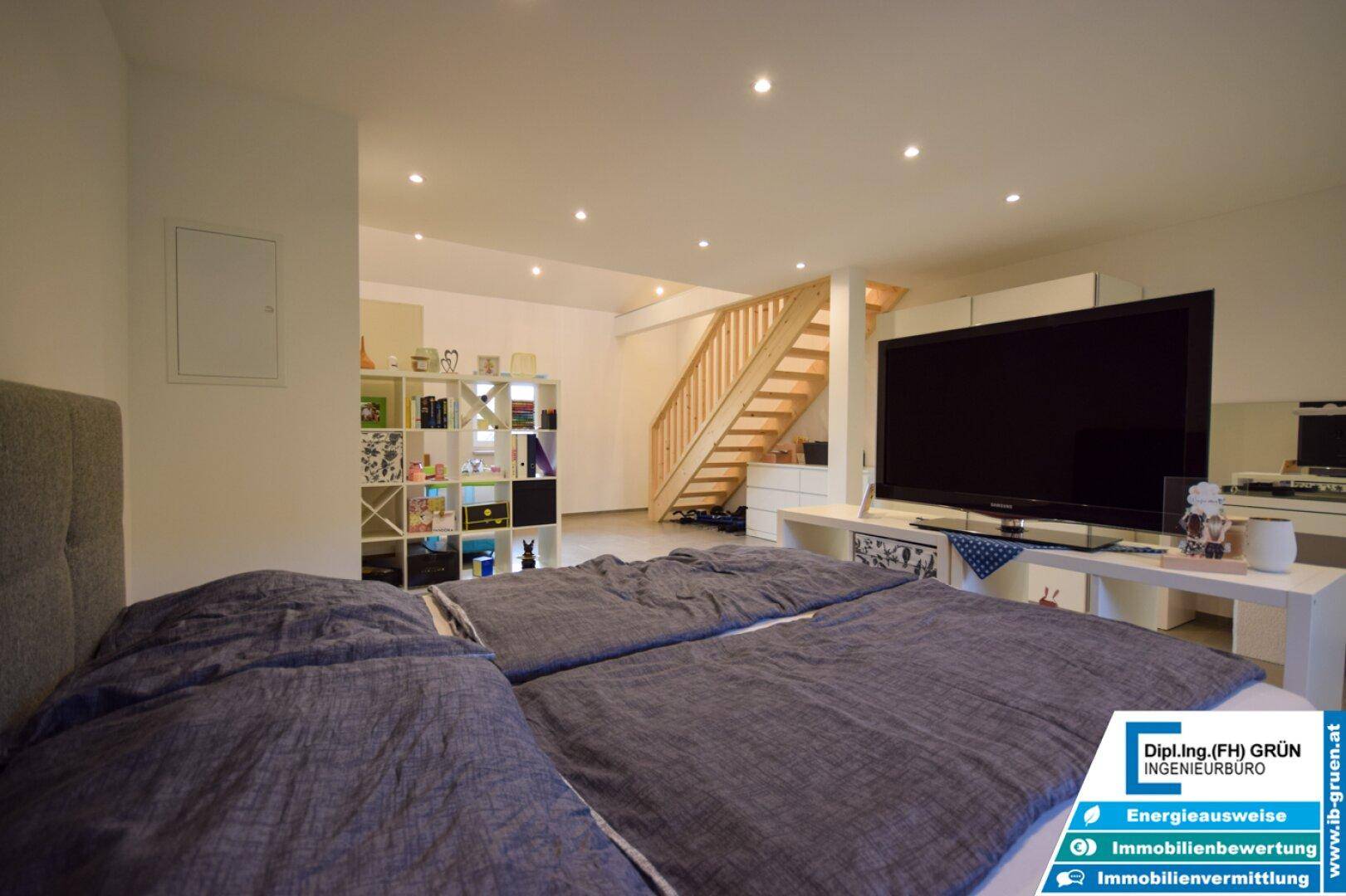 Geräumiges Schlafzimmer mit großem Fernseher und offener Holztreppe, die zu einer oberen Ebene führt.