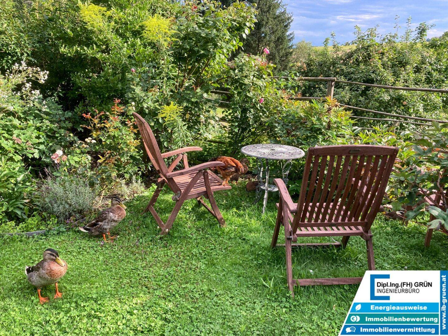 Gemütlicher Sitzbereich im Garten mit Holzstühlen und einem kleinen Tisch, umgeben von Pflanzen.
