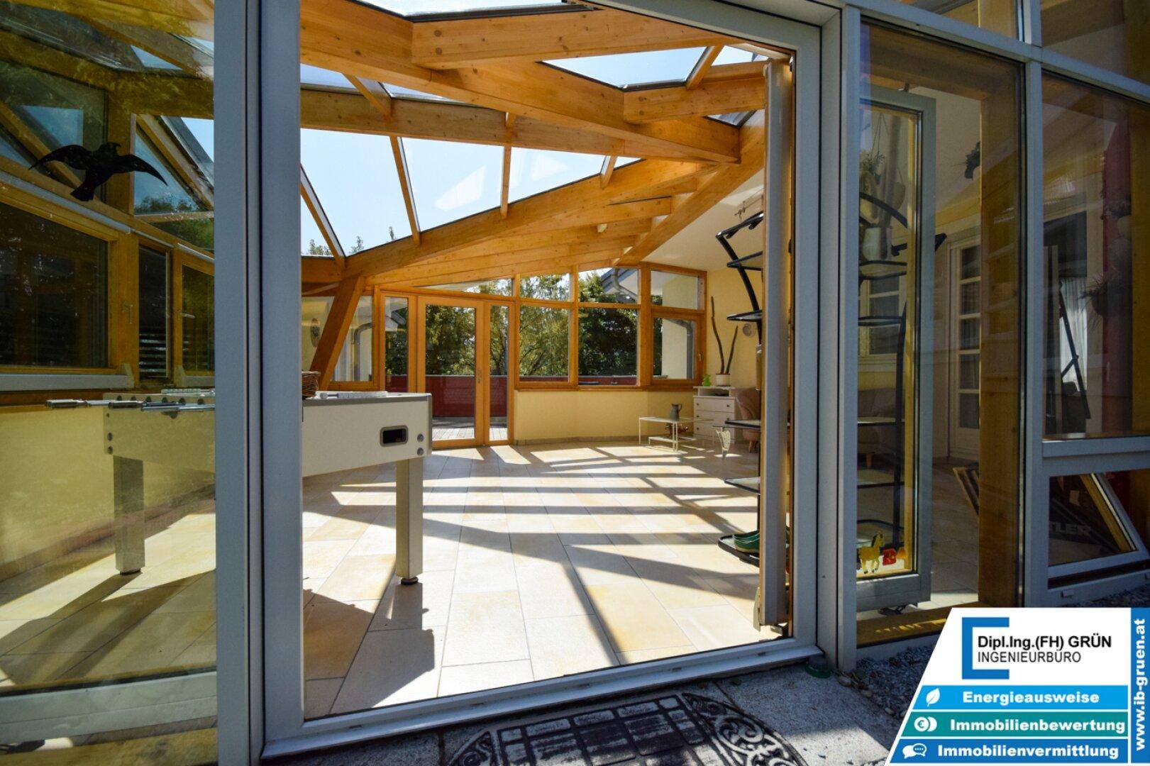 Blick von außen in den hellen Wintergarten mit Holzbalken und viel Glas.