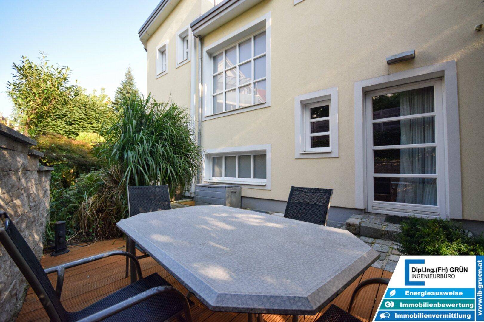 Terrasse mit Steintisch und Stühlen, umgeben von grüner Bepflanzung und dem Haus.