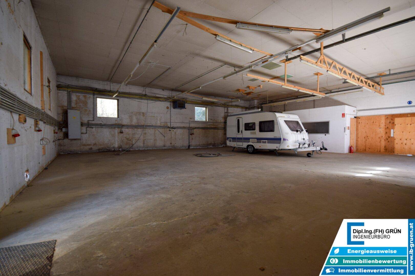 Großer, leerer Lagerraum mit Betonboden und einem geparkten Wohnwagen, beleuchtet von Deckenleuchten.
