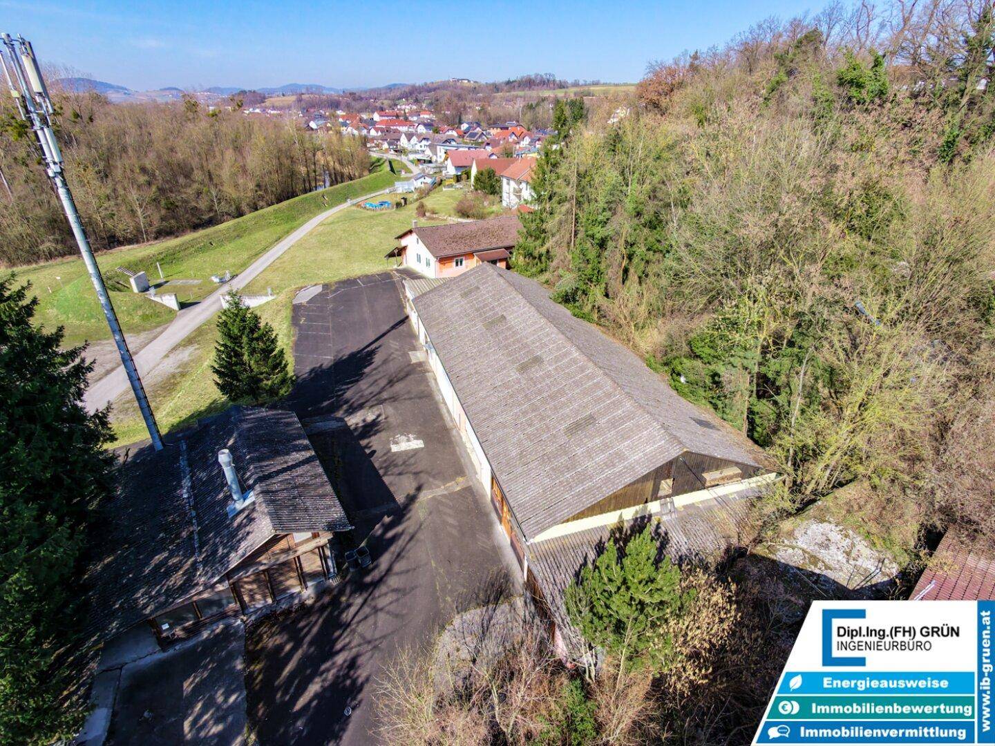 Vogelperspektive des Anwesens mit einem langen Gebäude und umliegenden Bäumen, im Hintergrund eine Siedlung.