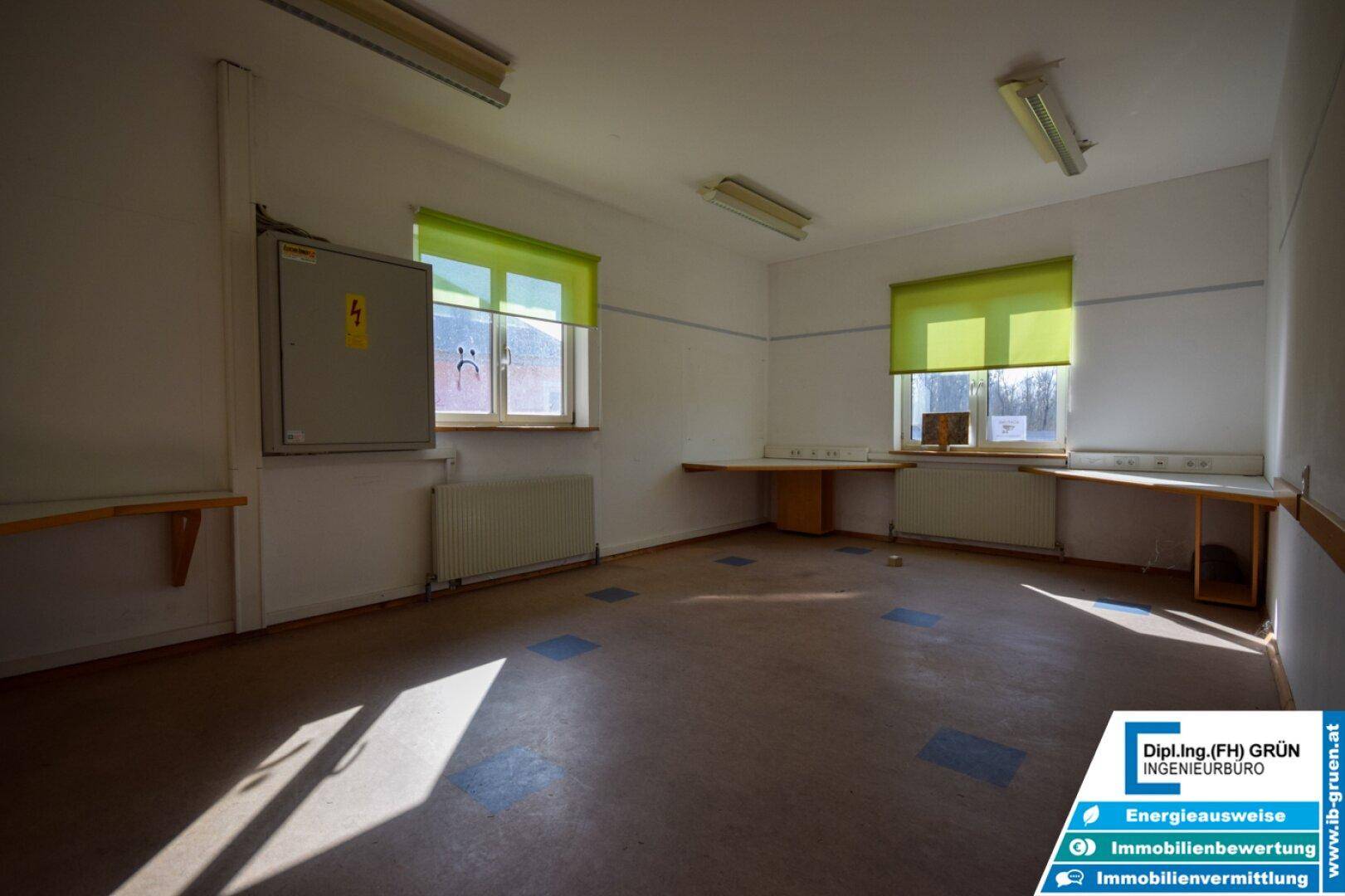 Heller Raum mit Linoleumboden, grünen Rollos an den Fenstern und Heizkörpern, möglicherweise ein Klassenzimmer.