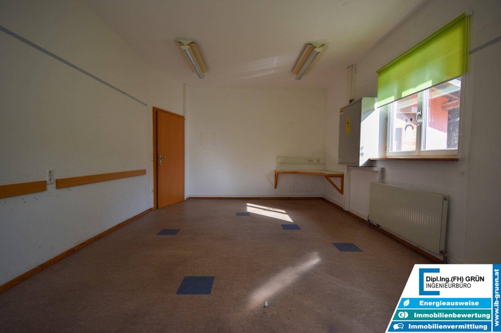 Raum mit Linoleumboden, einem Fenster mit grünem Rollo und einer Wandmontage für Elektrik.