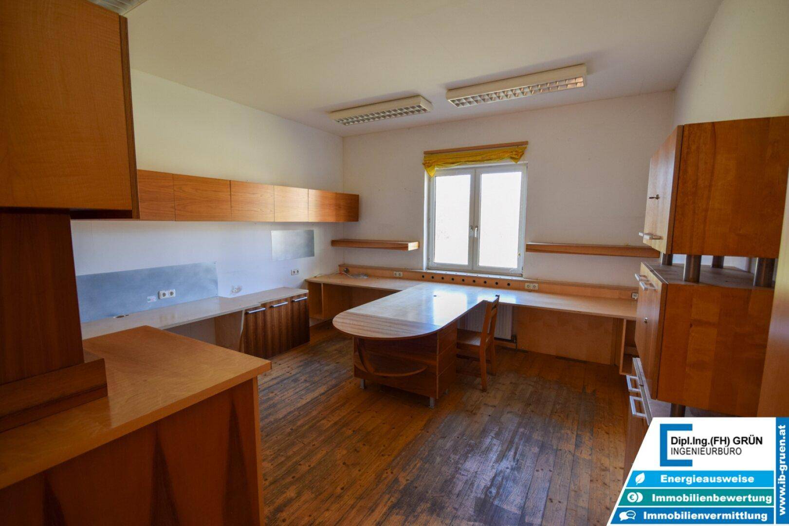 Großer Büroraum mit Holzdielenboden und umfangreichen Holzmöbeln, gut beleuchtet durch ein großes Fenster.