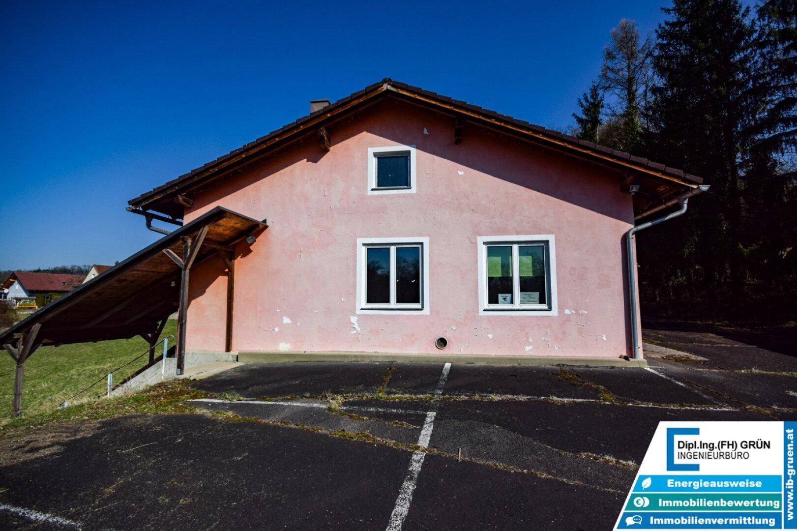 Rosa gestrichenes Gebäude mit Satteldach, Fenstern und einem Carport auf einem asphaltierten Parkplatz.