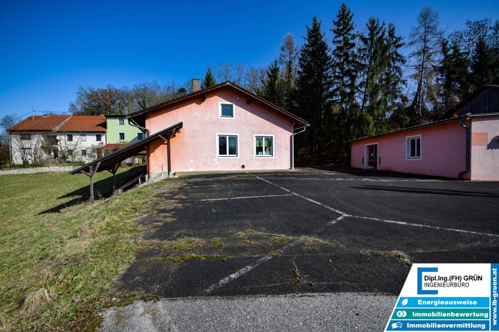 Gesamtansicht des rosa Gebäudes mit Carport und großem asphaltierten Parkplatz, umgeben von Bäumen.