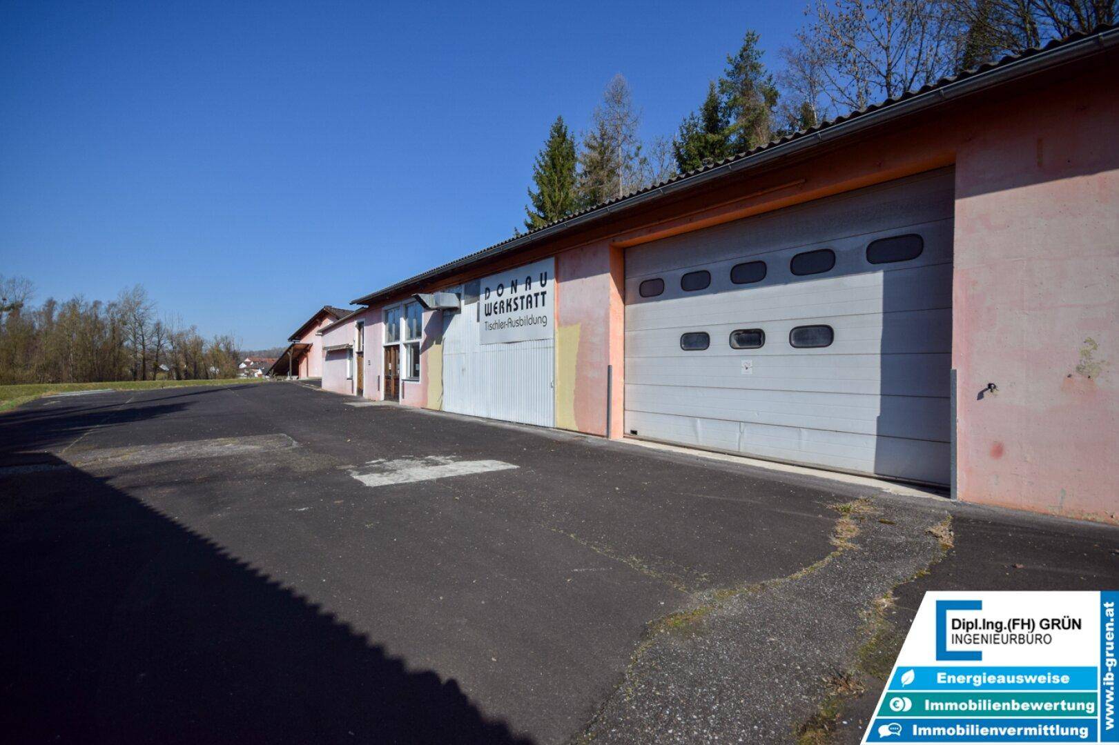 Lange Gebäudefassade mit Garagentoren und Fenstern, umgeben von einem asphaltierten Vorplatz.