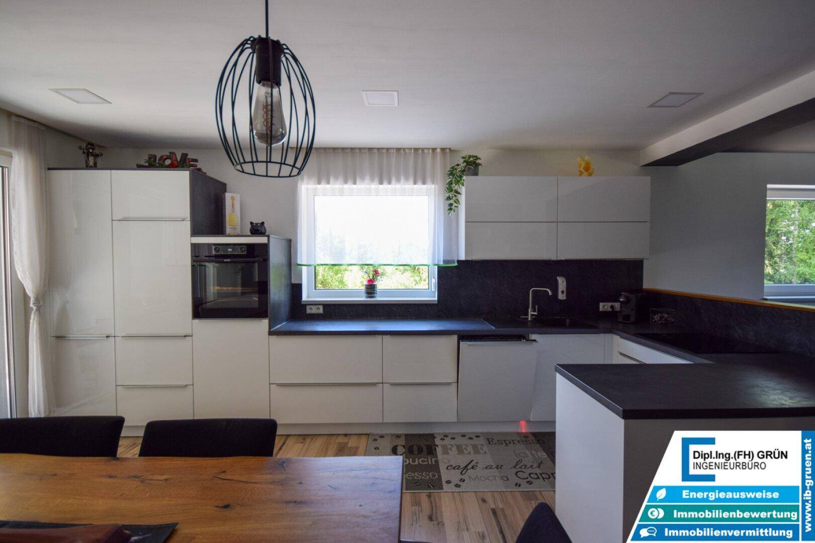 Moderne Küche mit weißen Schränken, dunklen Arbeitsplatten und integriertem Backofen am Fenster.