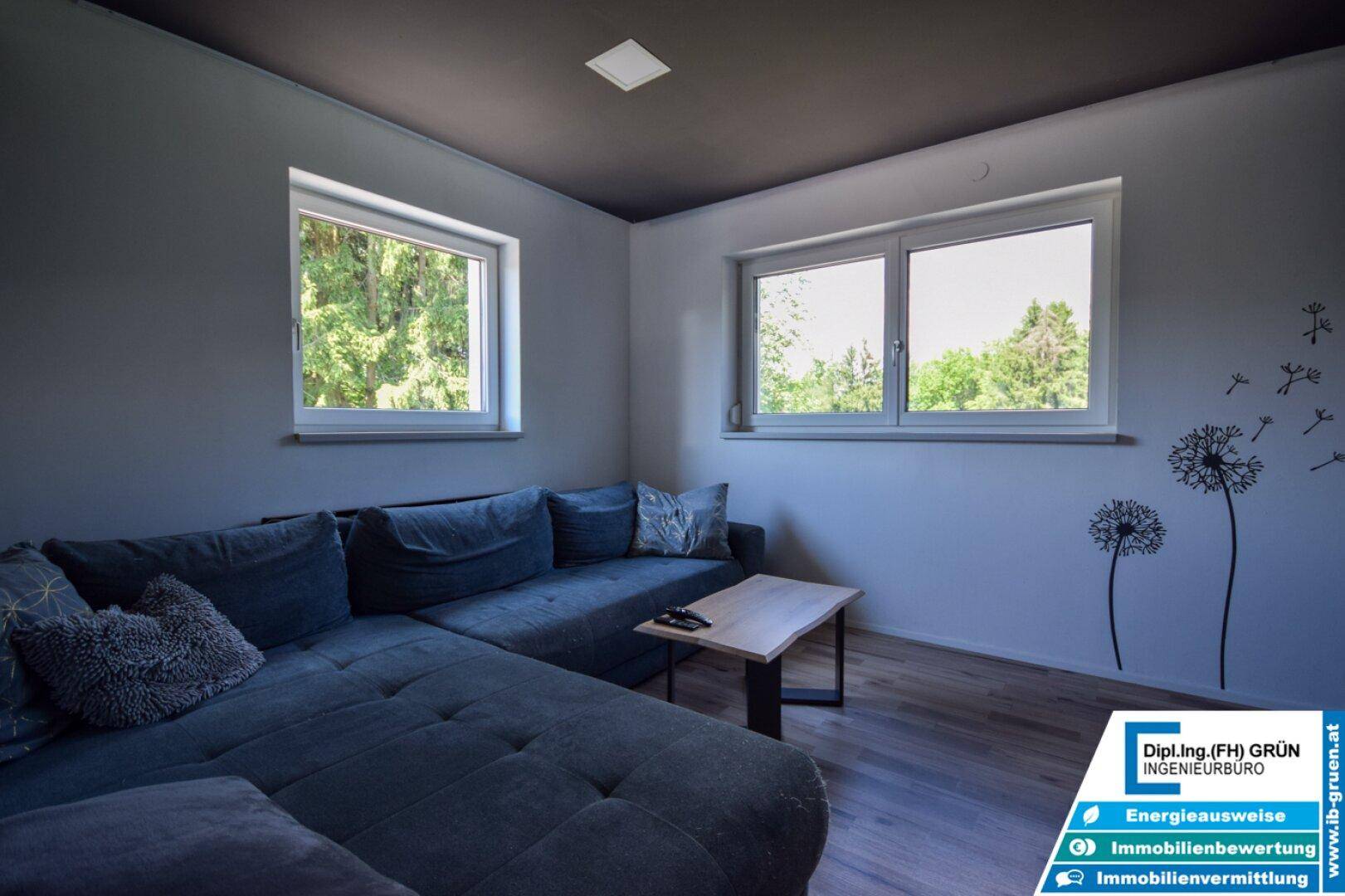 Gemütliches Wohnzimmer mit großem Sofa, Holztisch und Fenstern mit Blick ins Grüne.