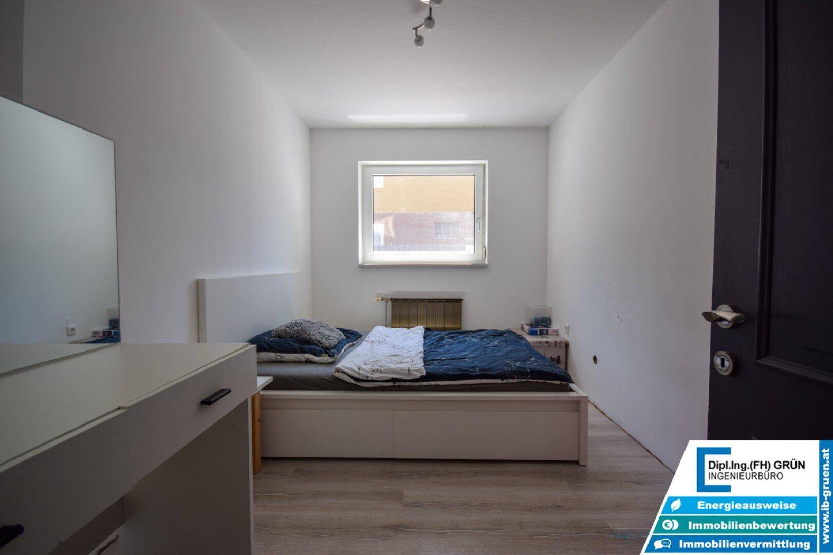 Helles Schlafzimmer mit einfachem Bett, weißen Wänden und einem Fenster für natürliches Licht.