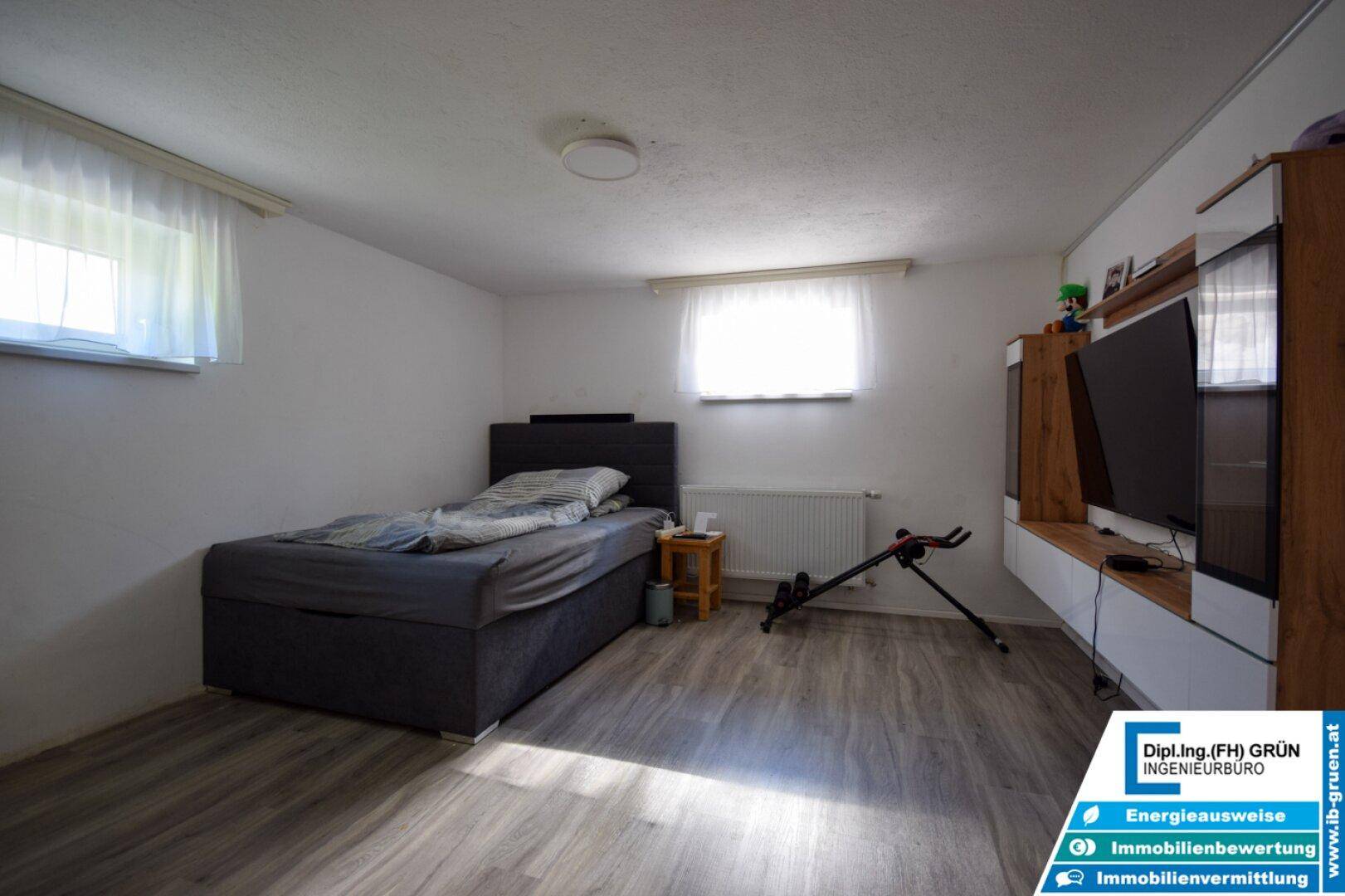 Geräumiges Schlafzimmer im Untergeschoss mit Bett, TV-Möbel und zwei Fenstern.