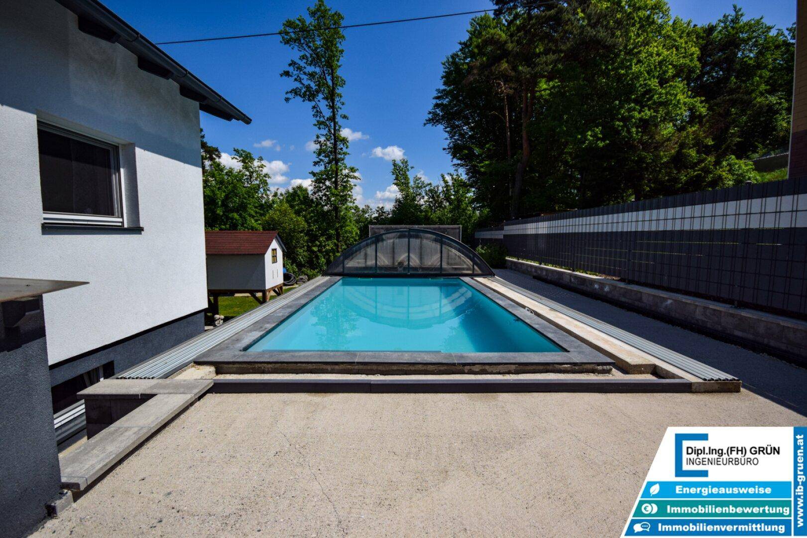 Überdachter Außenpool neben dem Haus, umgeben von viel Grün und einem sonnigen Himmel.