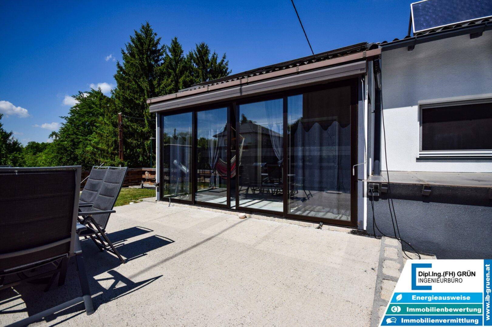 Sonnige Terrasse mit Liegestühlen und Blick auf den modernen Wintergarten mit großen Glasfronten.