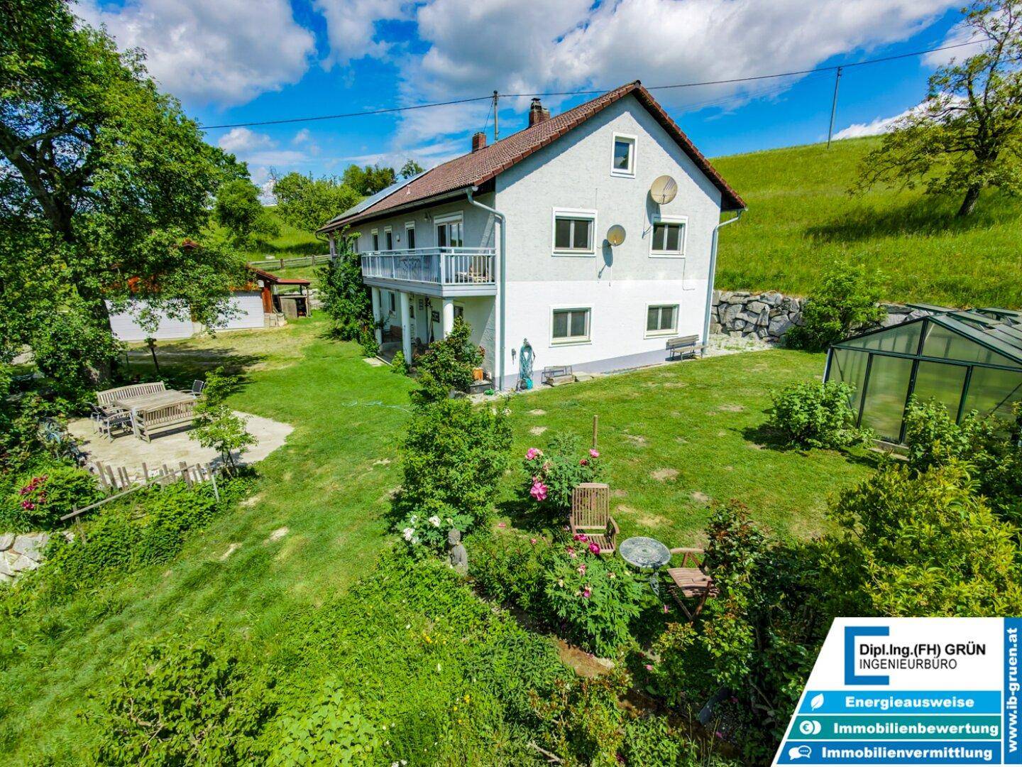 Hausfassade mit Balkon und weitläufigem Gartenbereich inklusive Gewächshaus und Sitzgelegenheiten.
