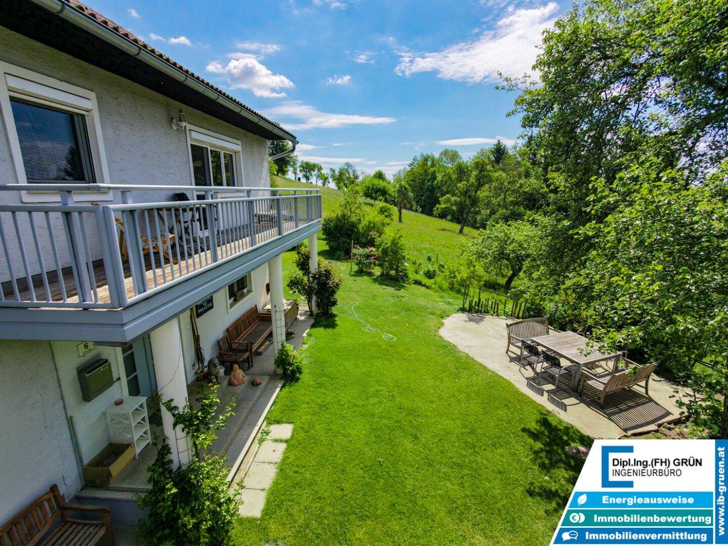 Seitlicher Blick auf das Haus mit Balkon, großer Rasenfläche und gepflasterter Terrasse mit Esstisch.