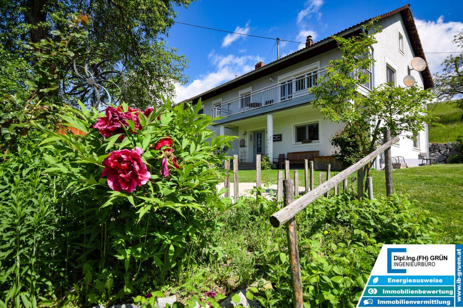 Hausfassade mit Balkon und üppigem Gartenbereich, geschmückt mit blühenden Pfingstrosen.