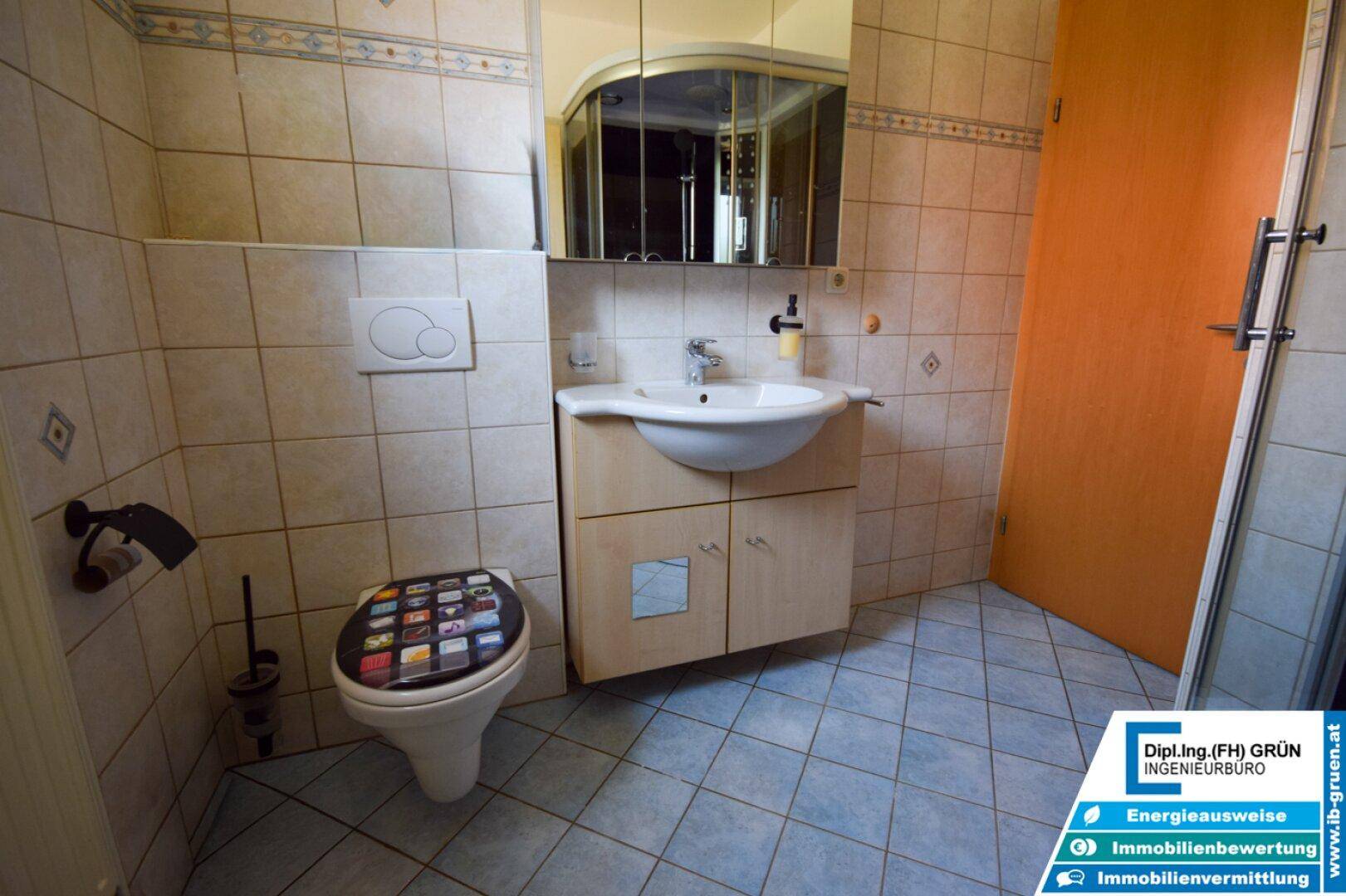 Gefliestes Badezimmer mit Waschbecken, Toilette und Spiegelschrank, ausgestattet mit einem farbigen Toilettendeckel.