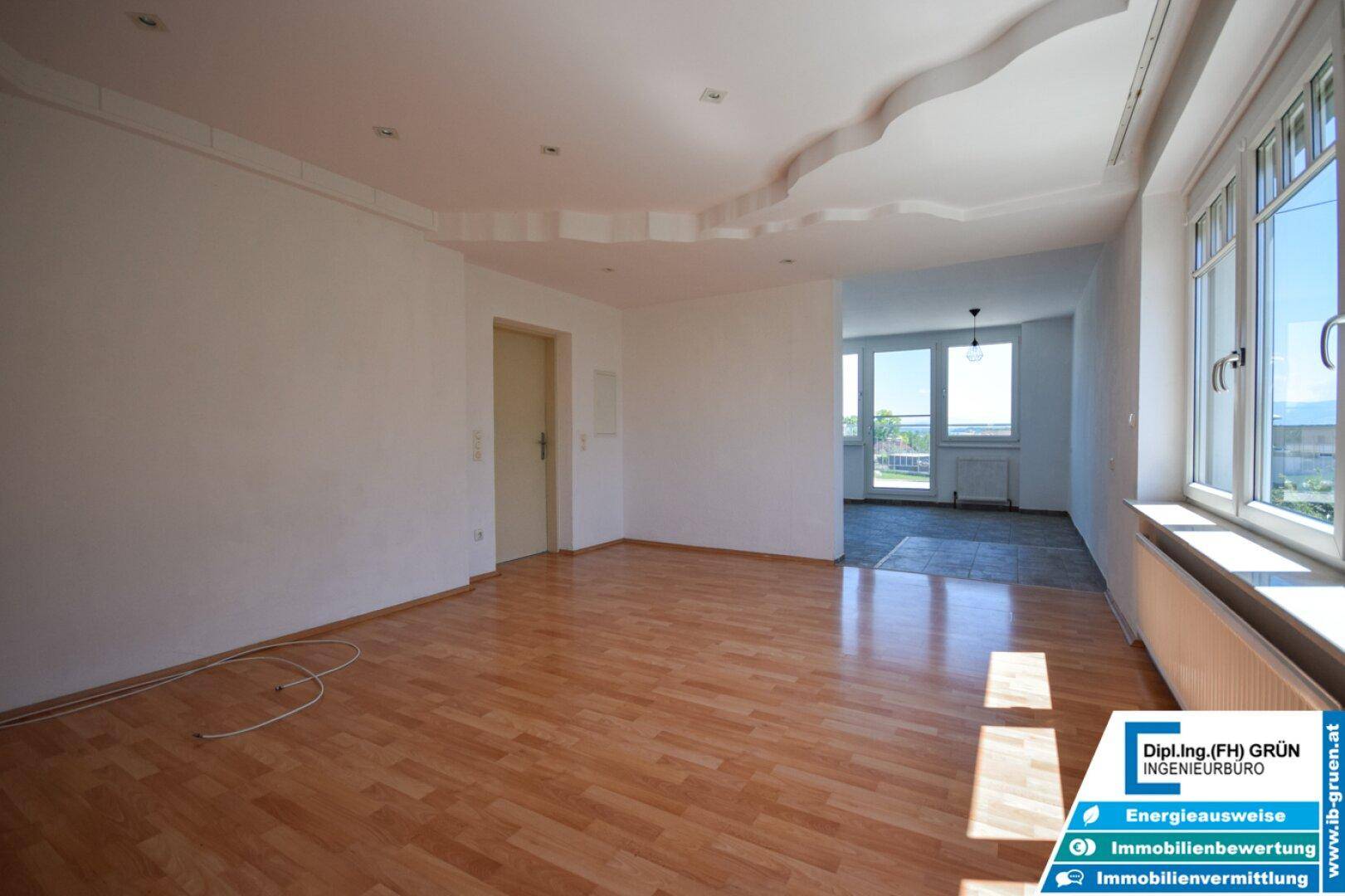 Geräumiges, leeres Wohnzimmer mit Holzboden, großen Fenstern und Zugang zu einem weiteren Raum.