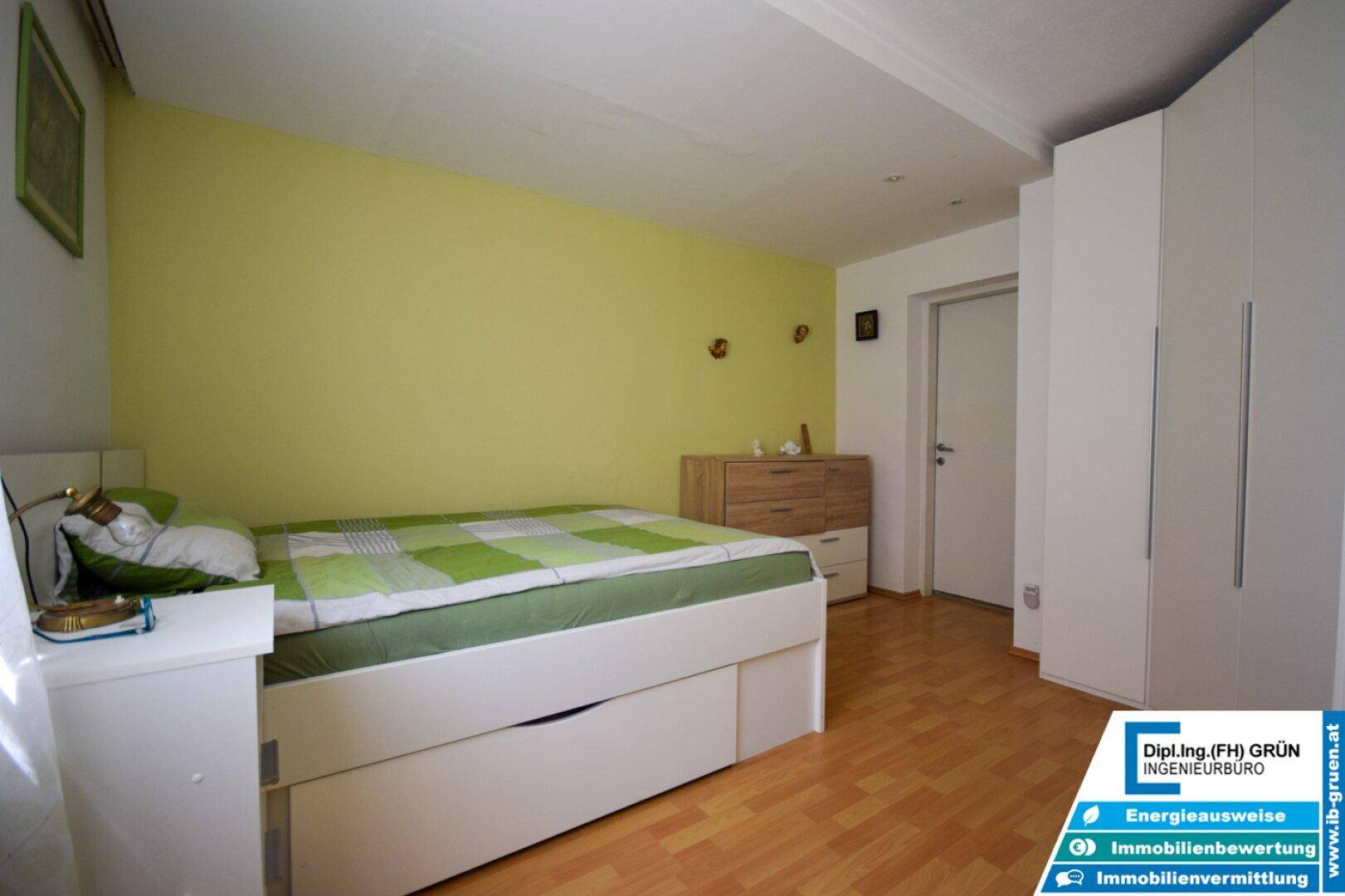 Schlafzimmer mit grün gestrichener Wand, Holzboden und weißem Kleiderschrank.