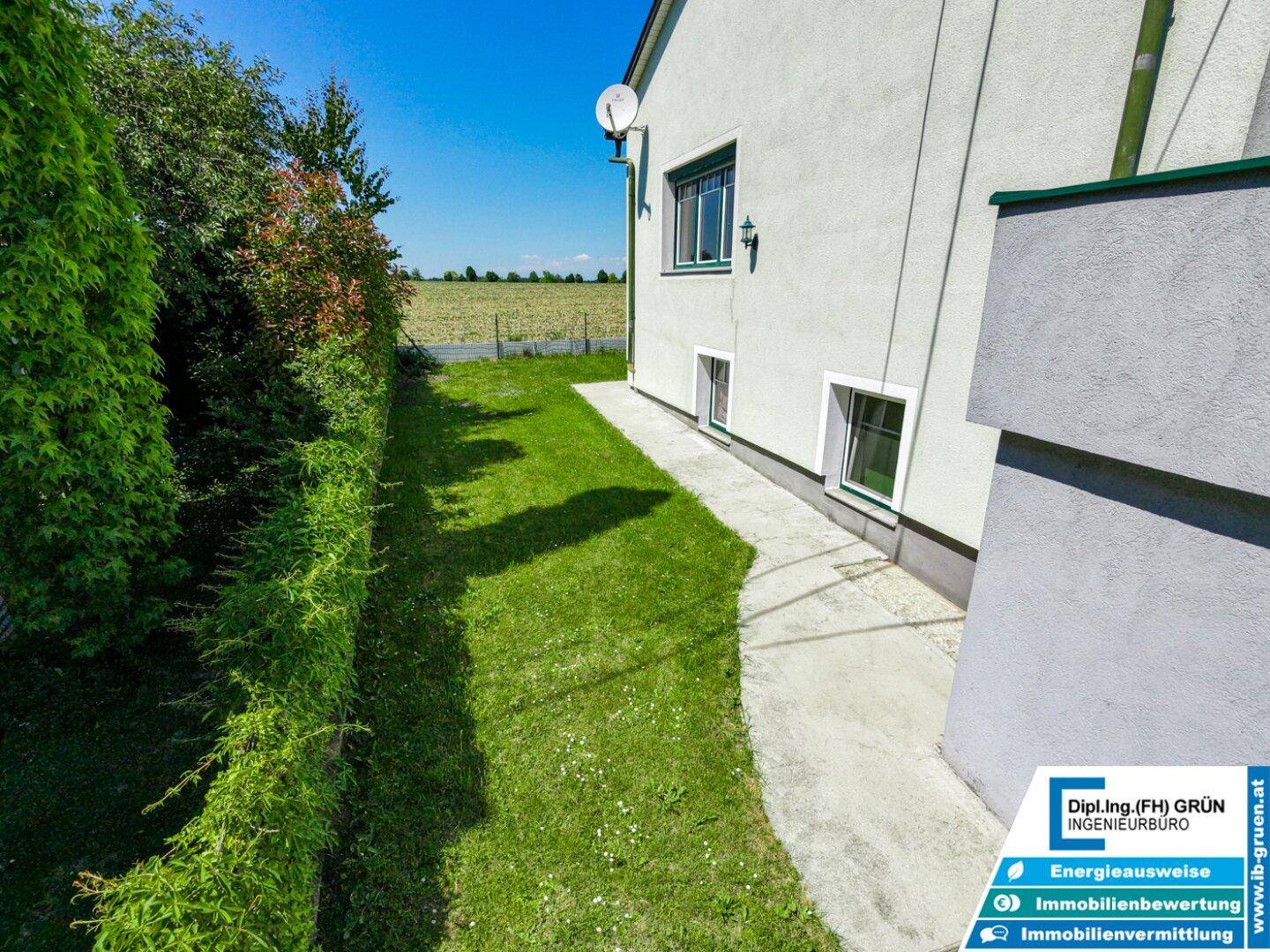 Seitenansicht des Hauses mit gepflegtem Rasen, Hecke und weitem Blick ins Grüne.