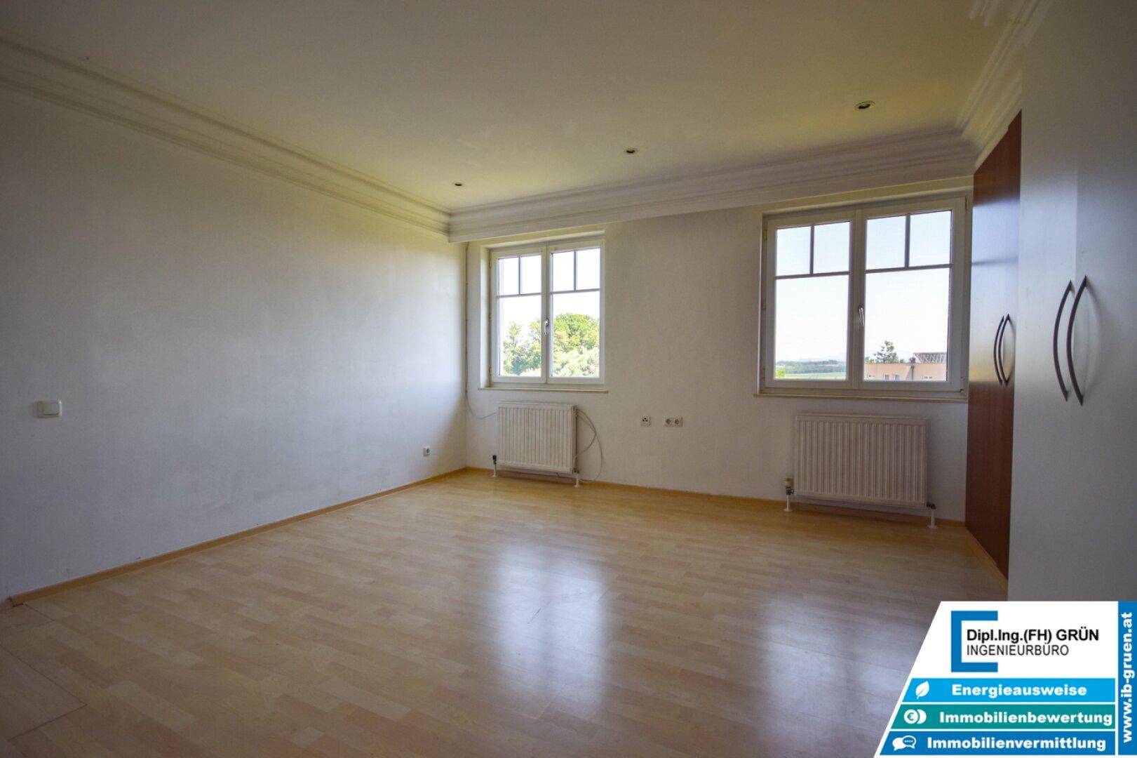 Geräumiges, helles Zimmer mit Laminatboden, zwei Fenstern und klassischer Deckenleiste.