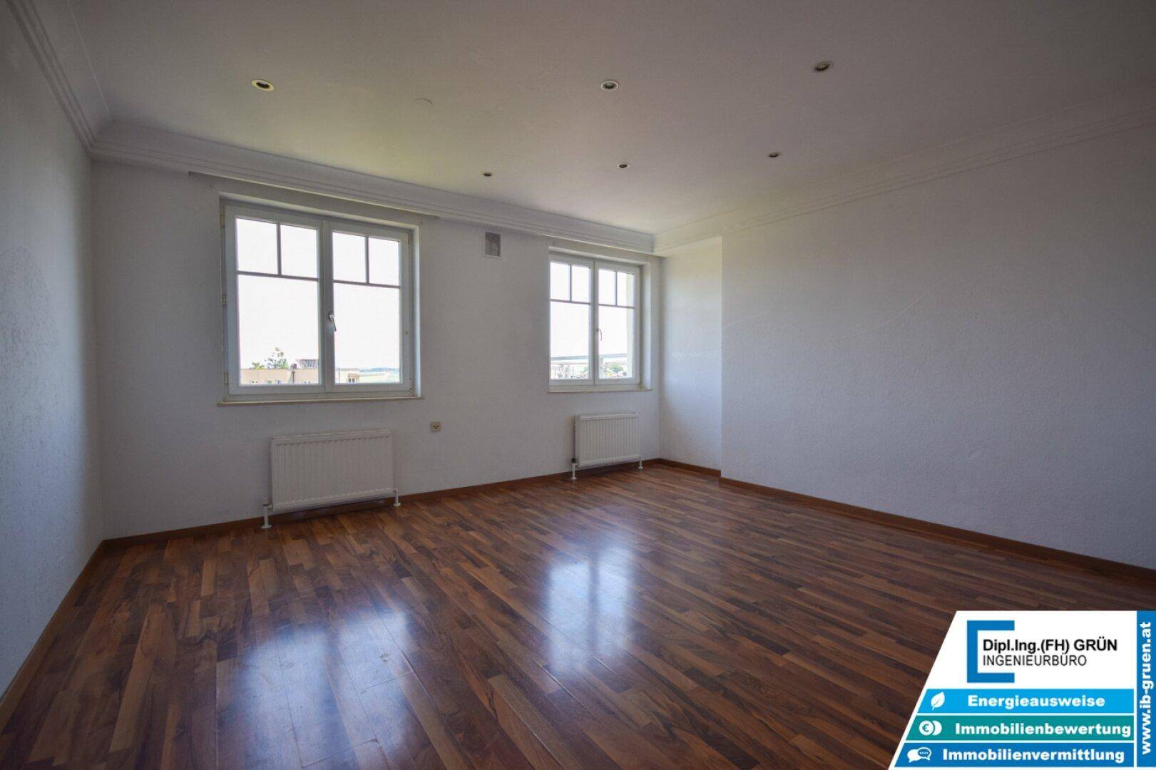 Großes, helles Zimmer mit dunklem Laminatboden, zwei Fenstern und eleganter Deckenleiste.