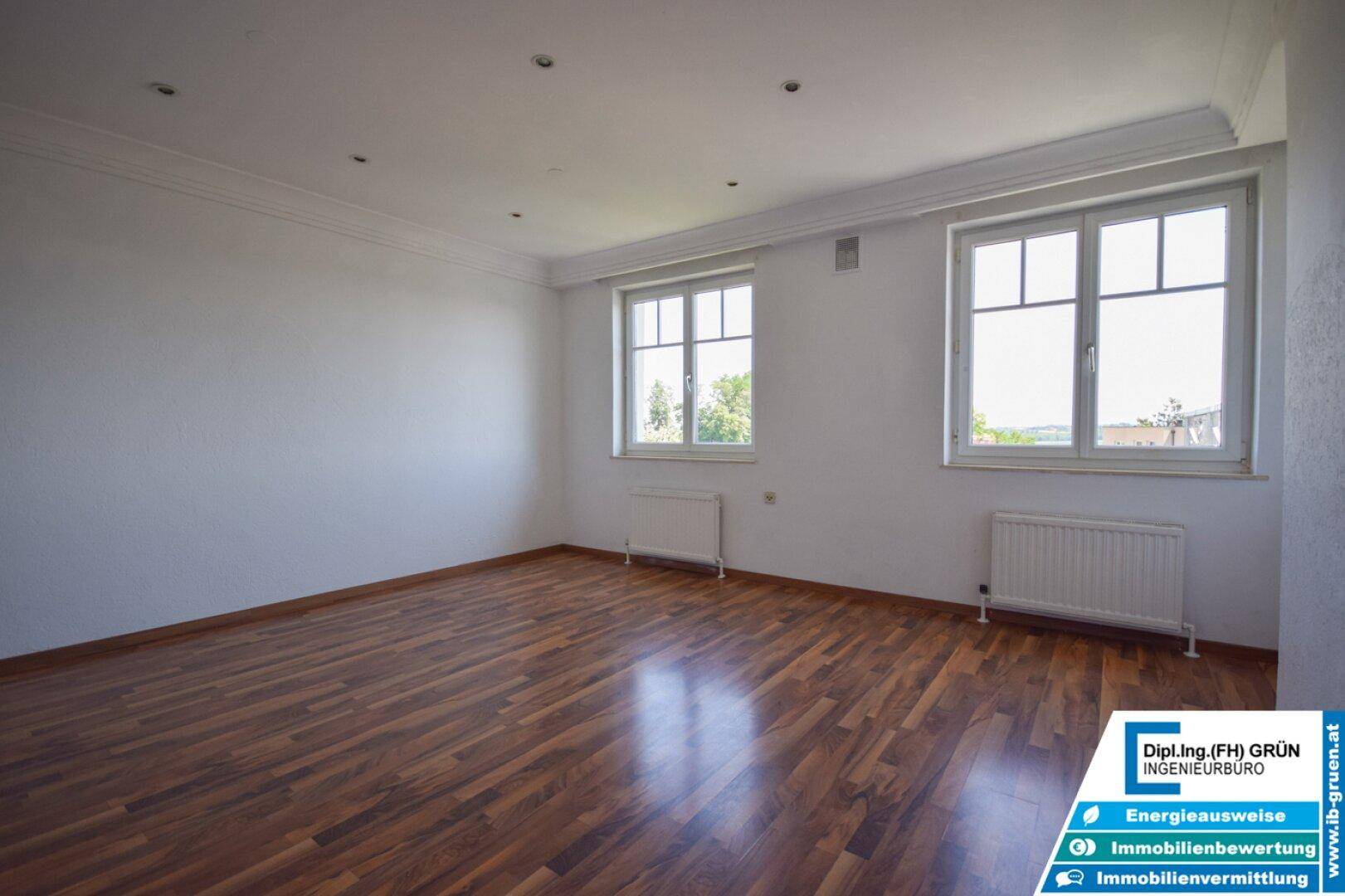 Helles Wohnzimmer mit dunklem Laminatboden und zwei großen Fenstern für viel Tageslicht.