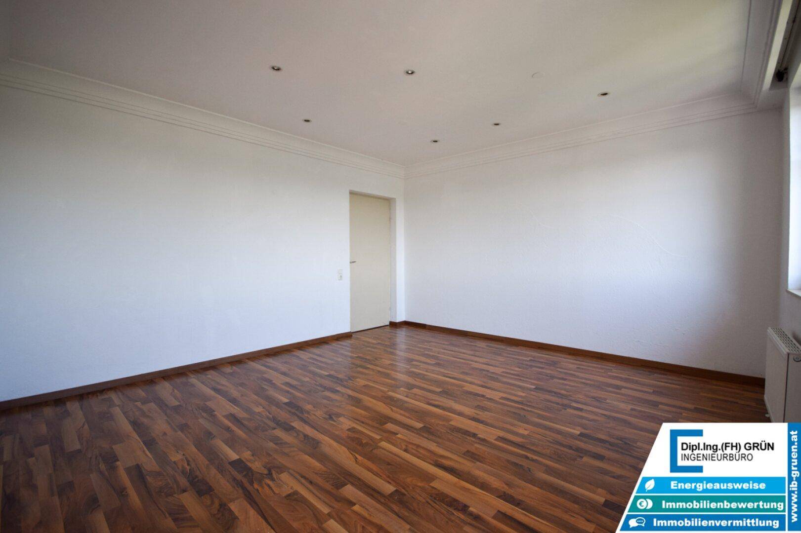 Geräumiges Zimmer mit dunklem Laminatboden und weiß gestrichenen Wänden, bietet viel Platz.