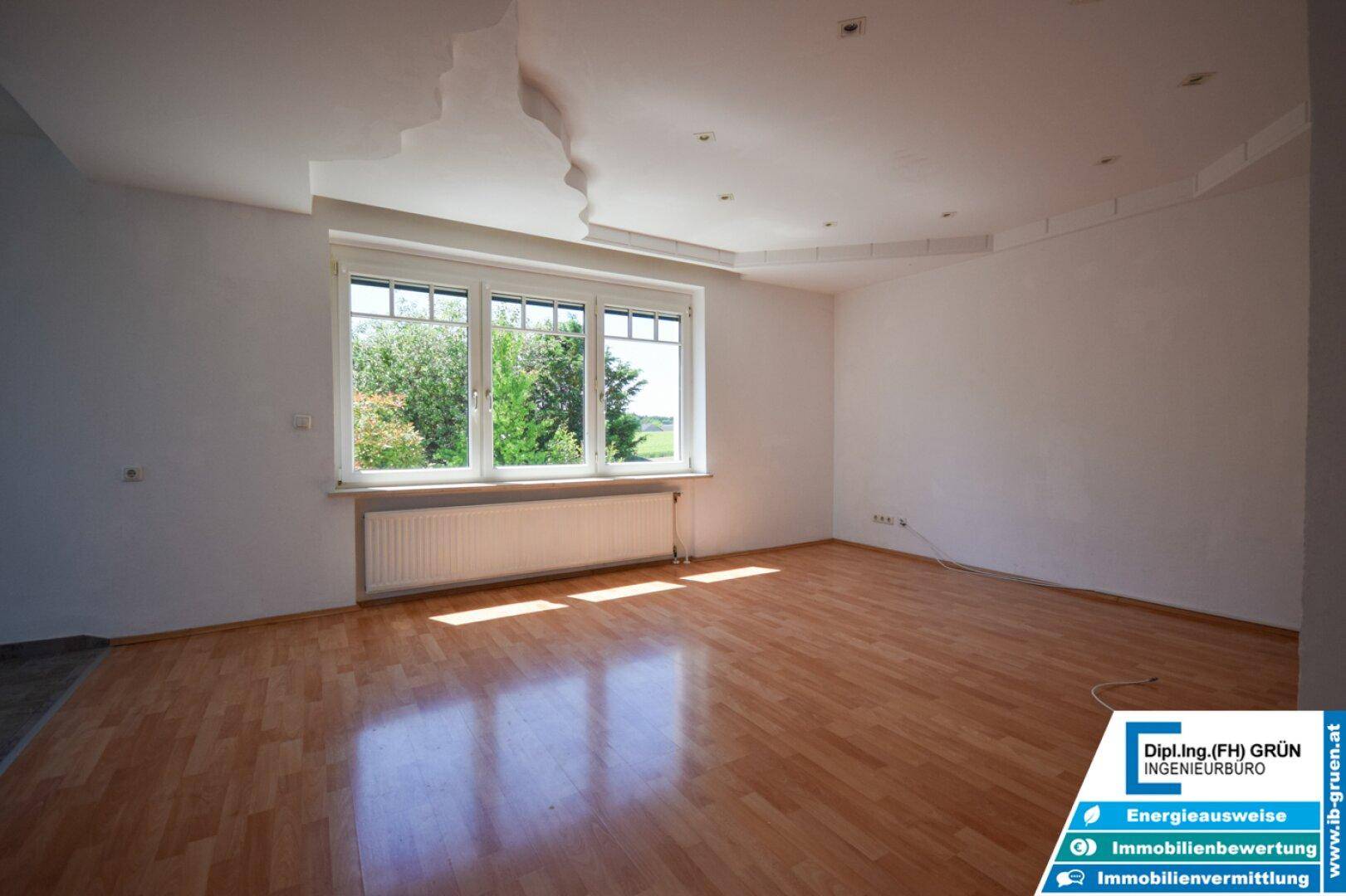 Helles Wohnzimmer mit Laminatboden, großem Fenster und dekorativer Deckenstruktur.
