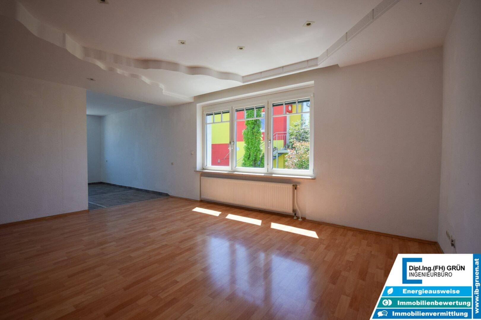 Helles Wohnzimmer mit Laminatboden und großem Fenster, das viel Tageslicht hereinlässt.