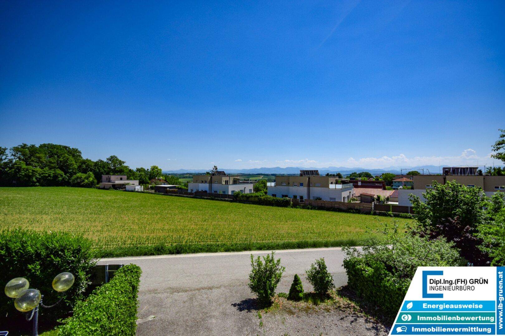 Weitläufiger Blick über grüne Felder und die umliegende Landschaft mit Bergen am Horizont.