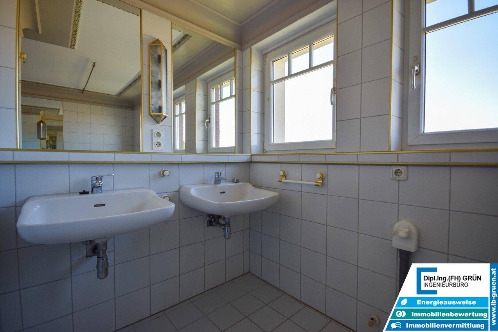Badezimmer mit zwei Waschbecken, großen Spiegeln und weißen Fliesen mit goldenen Akzenten.