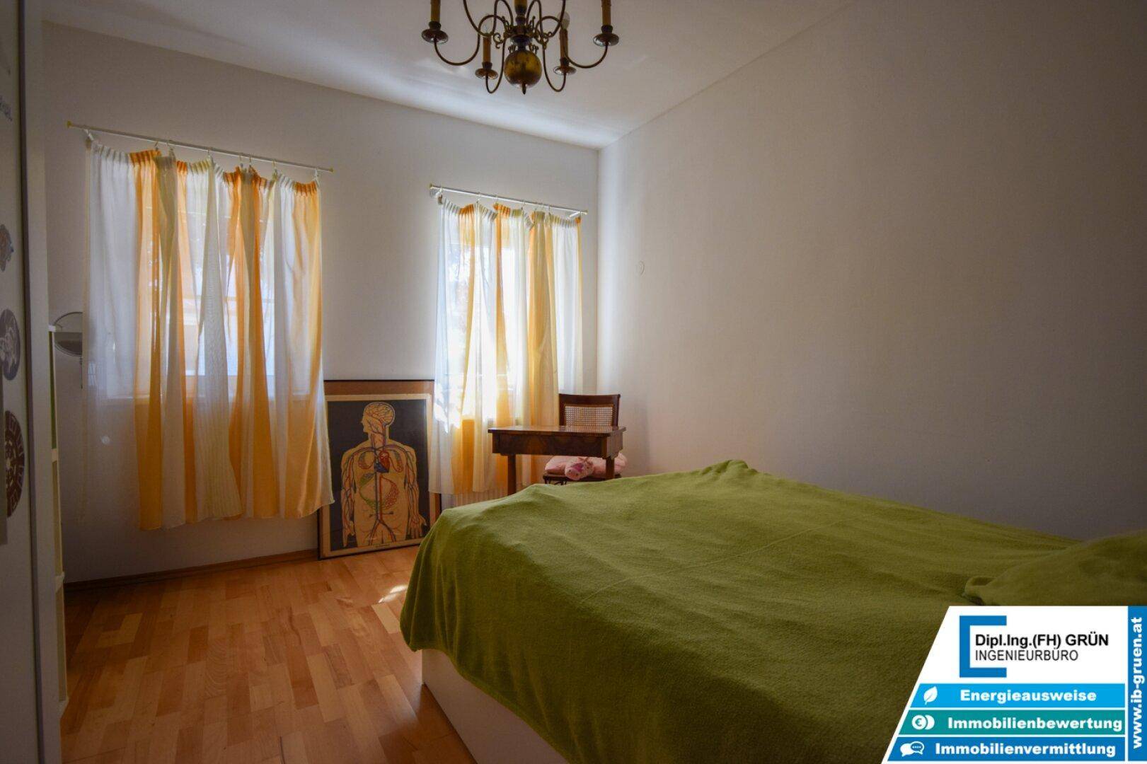 Schlafzimmer mit Parkettboden, zwei Fenstern und einem Bett mit grüner Decke.