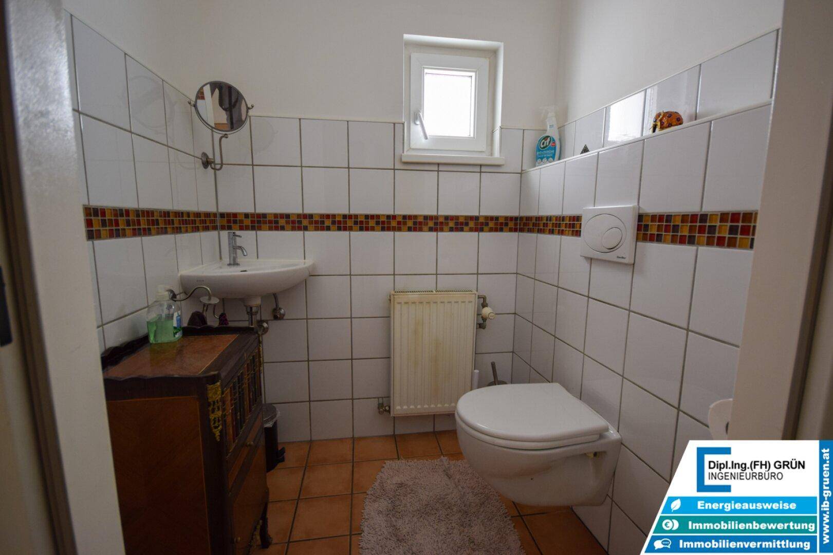 Kleines WC mit Fenster, weißen Fliesen und Terrakotta-Bodenfliesen, ausgestattet mit einem Waschbecken.
