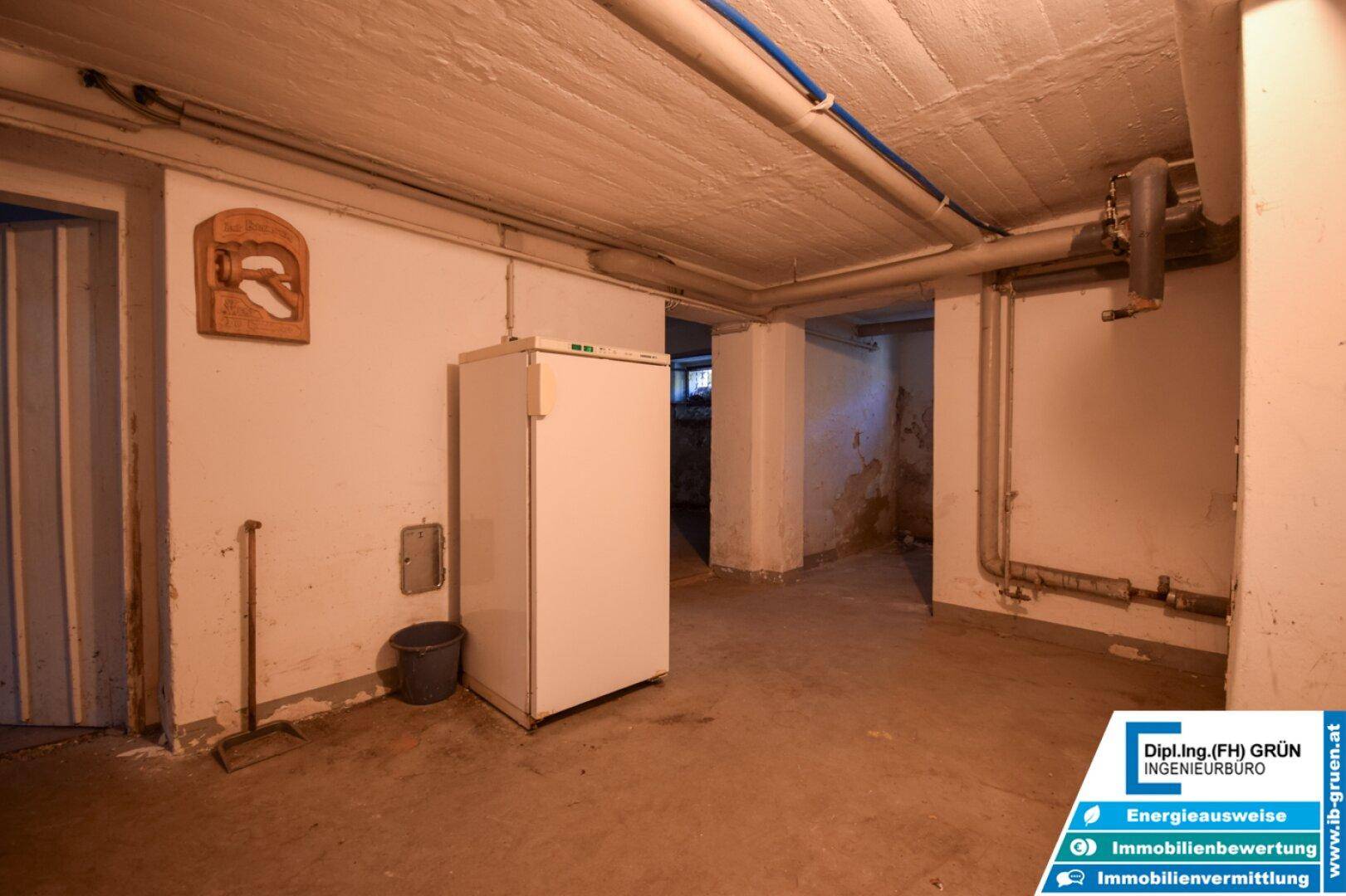 Geräumiger Kellerraum mit Betonboden, freistehendem Kühlschrank und sichtbaren Rohren.