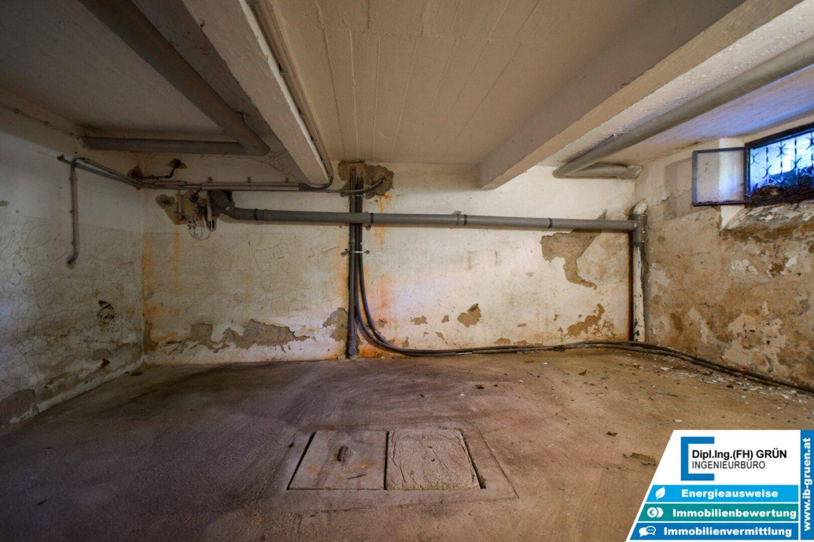 Unausgebauter Kellerraum mit sichtbaren Rohren, Betonboden und einem kleinen Fenster.