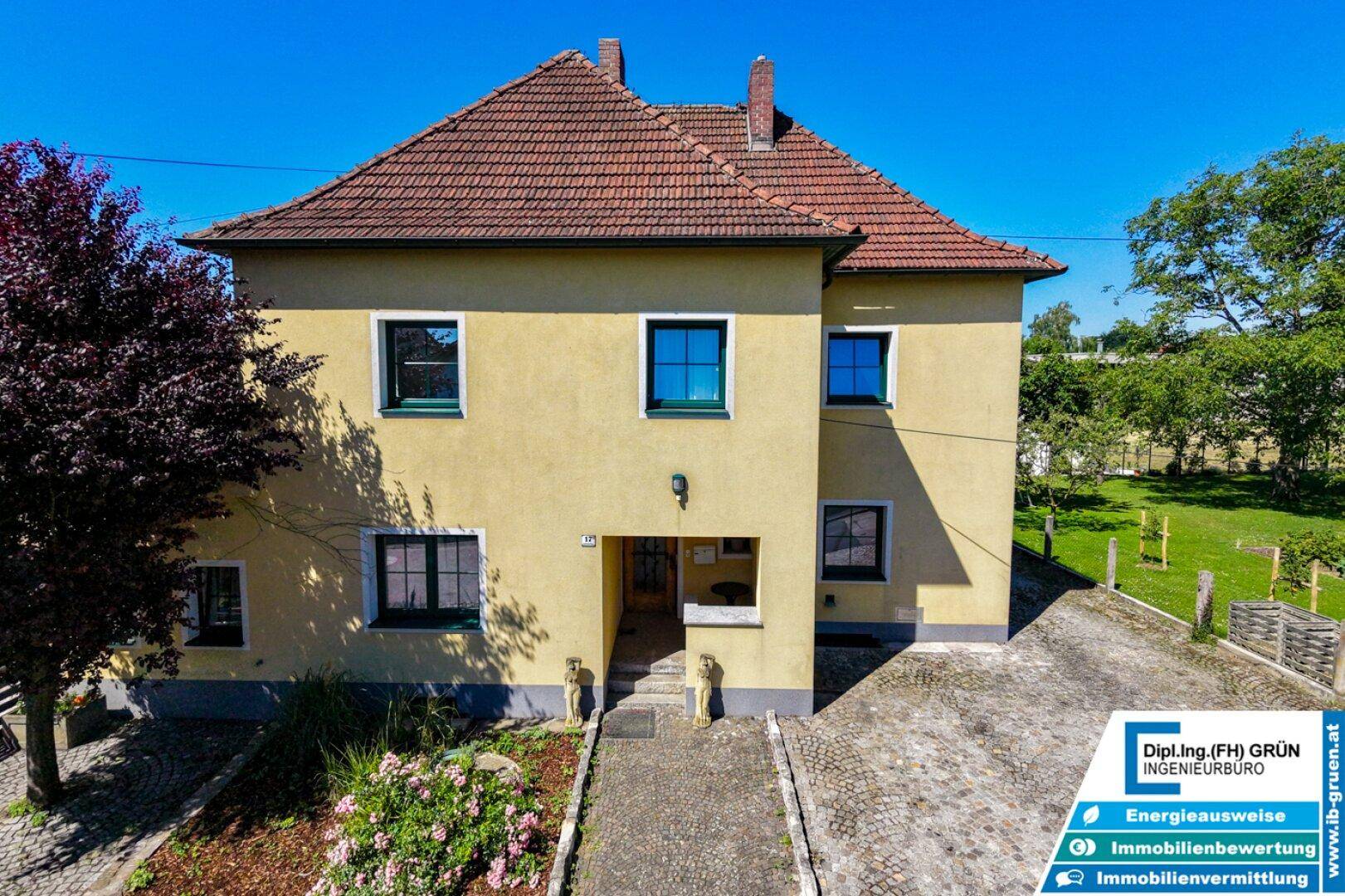 Ansicht des Hauses von vorne mit gelber Fassade, roten Dachziegeln und Eingangsbereich.