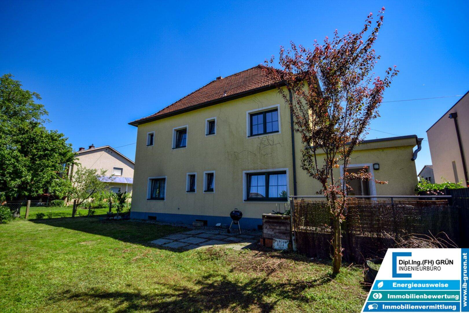 Gelbes Einfamilienhaus mit großem Garten und gepflasterter Terrasse unter blauem Himmel.