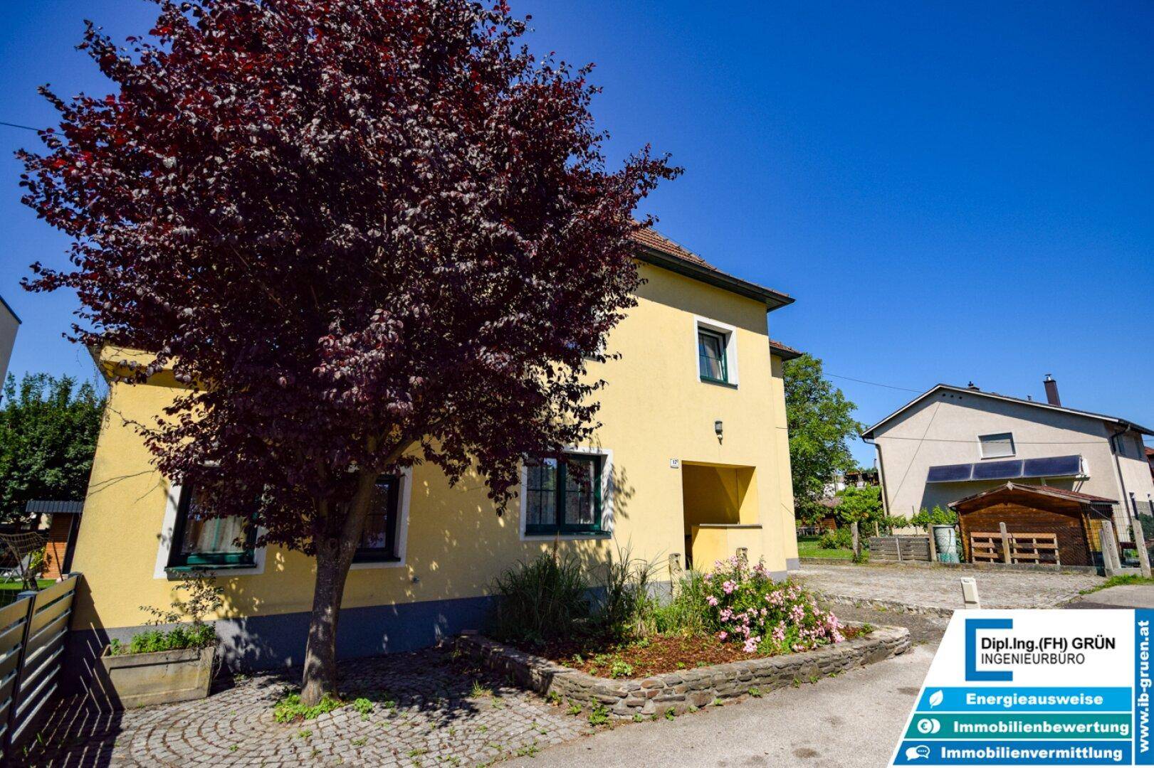 Gelbes Wohnhaus mit rotem Baum im Vorgarten und gepflastertem Zugangsbereich.