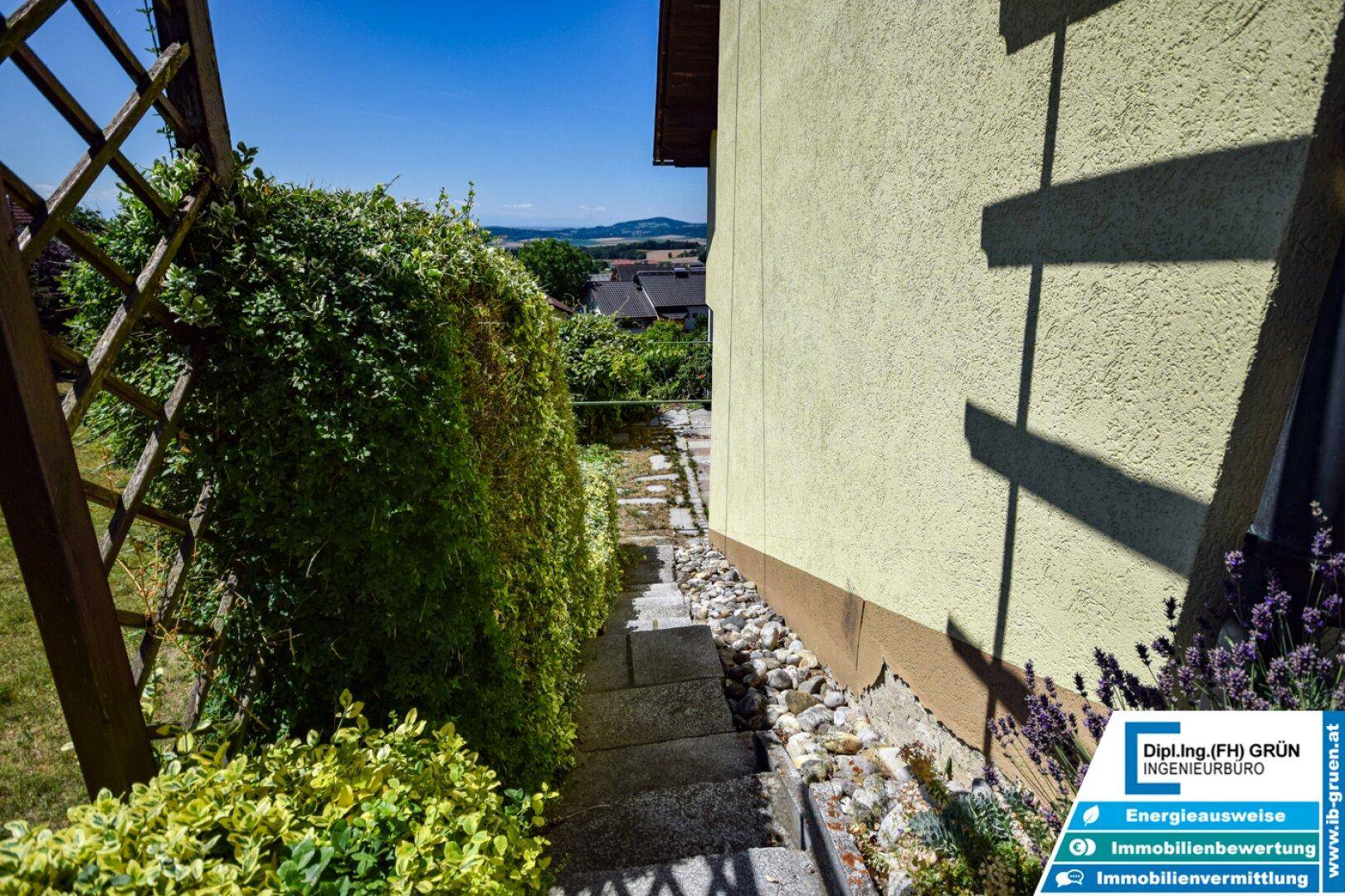Seitlicher Gartenweg mit üppigem Grün und Steinstufen, führt um das Haus herum.