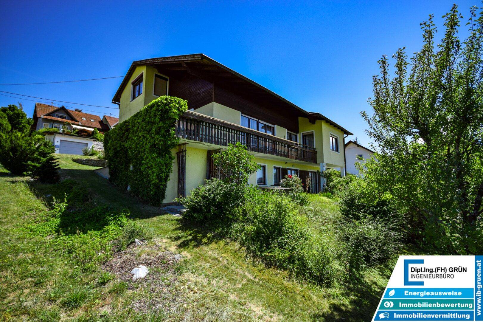 Hausfassade mit Balkon und üppiger Vegetation in einer idyllischen, grünen Umgebung.