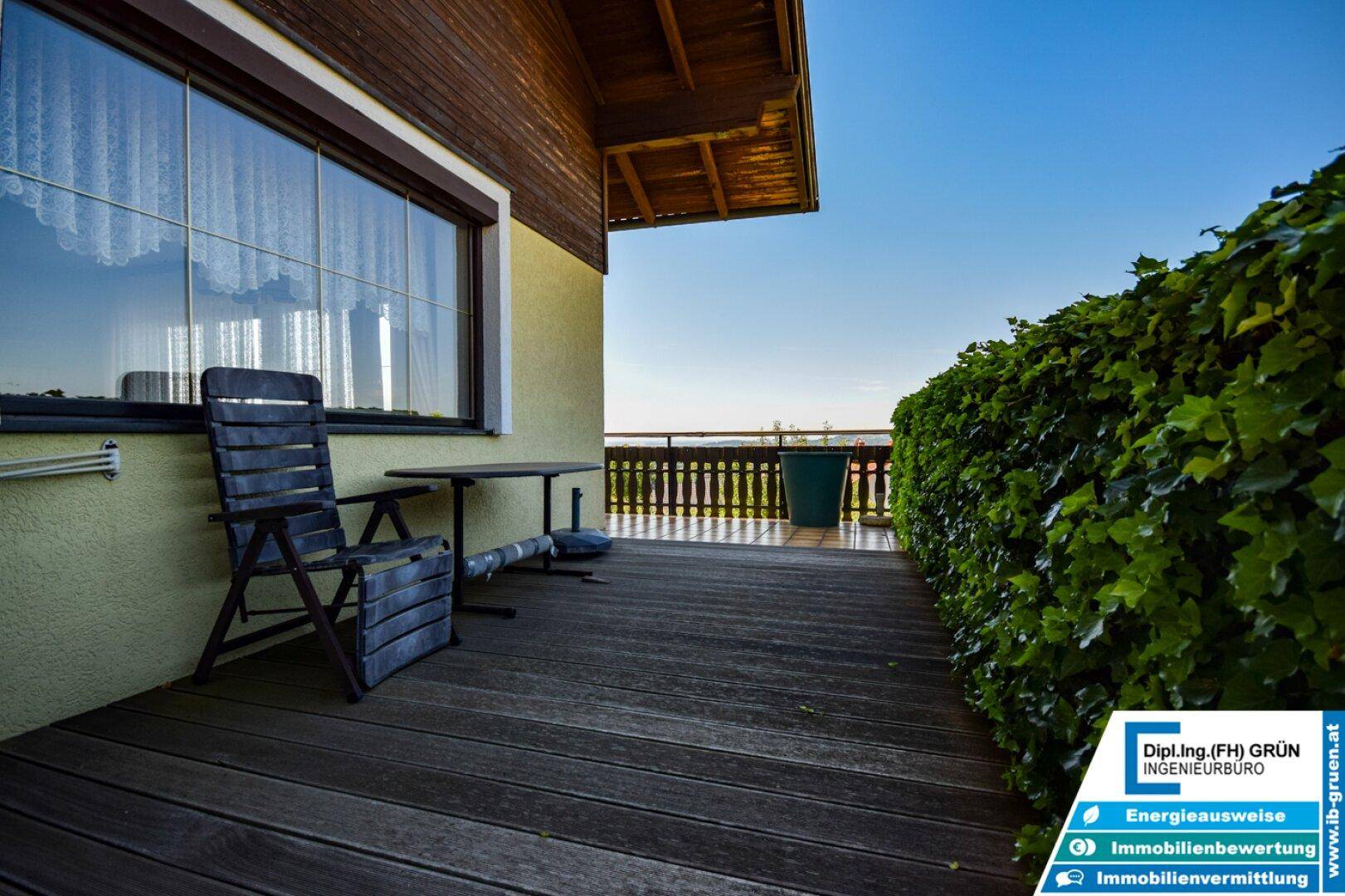 Balkon mit Holzboden und Sitzgelegenheit, umgeben von grünen Pflanzen und Weitblick.