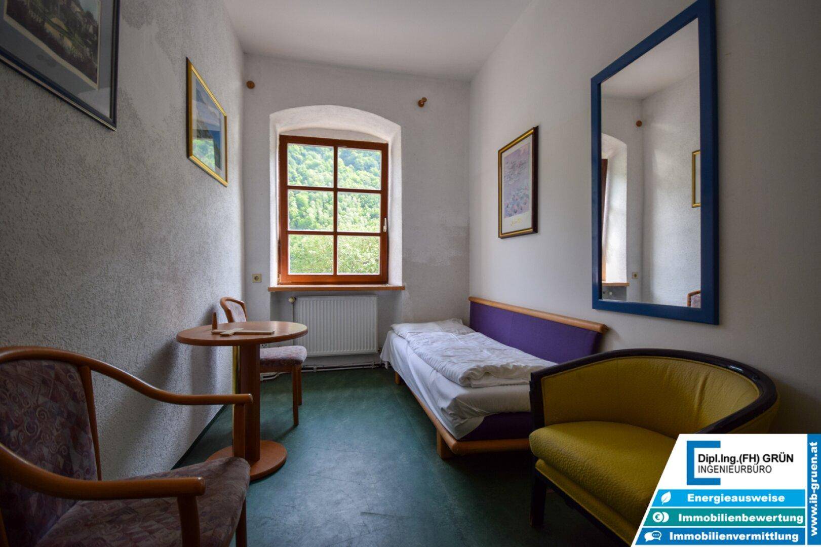Einfaches Schlafzimmer mit Einzelbett, kleinem Tisch und Fenster mit Grünblick.