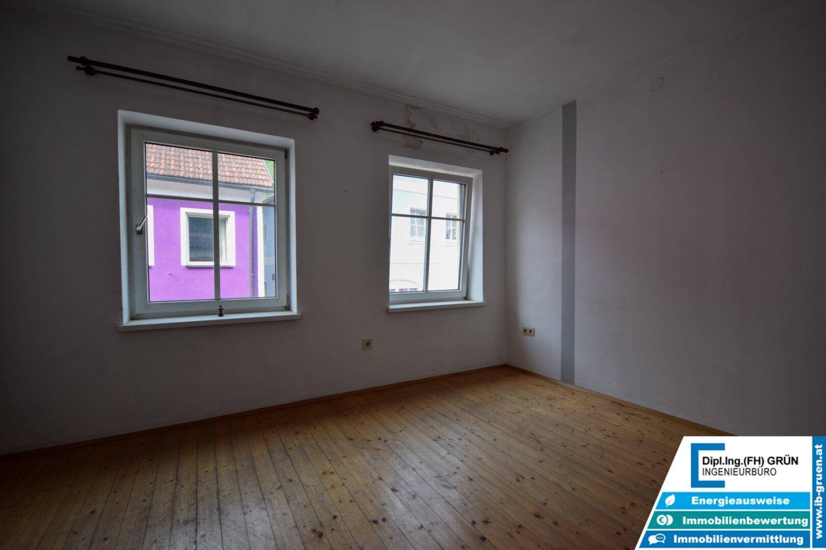Helles Schlafzimmer mit Holzdielenboden und zwei Fenstern mit Blick nach draußen.