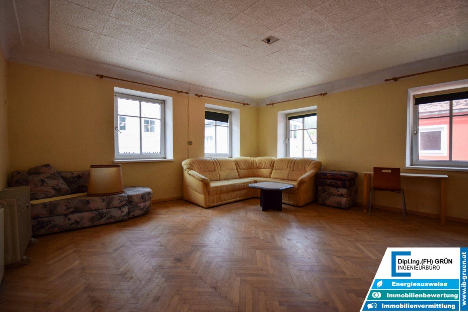 Geräumiges Wohnzimmer mit Fischgrätparkett, mehreren Fenstern und älteren Möbeln in traditionellem Stil.